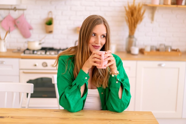 Pretty blonde woman drinking coffe tea on her kitchen, bright syulish loungewear.