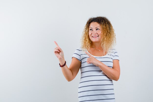 Pretty blonde woman pointing at upper left corner in striped t-shirt and looking merry. front view.