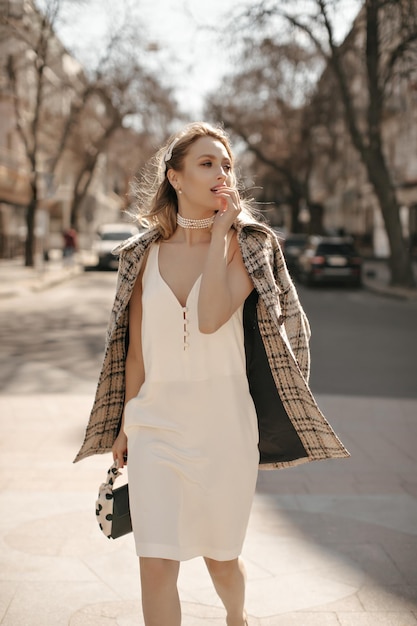 Free photo pretty blonde woman in tweed checkered jacket and silk white dress walking outside attractive lady in pearl necklace holding black handbag