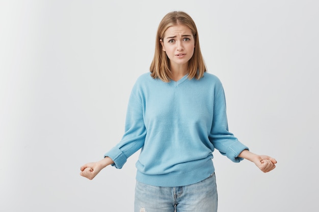 Free Photo pretty blonde young female with dark eyes,frowning her eyebrows while looking st camera with upset and thoughtful look. puzzled and dissatisfied cute girl in blue sweater and jeans