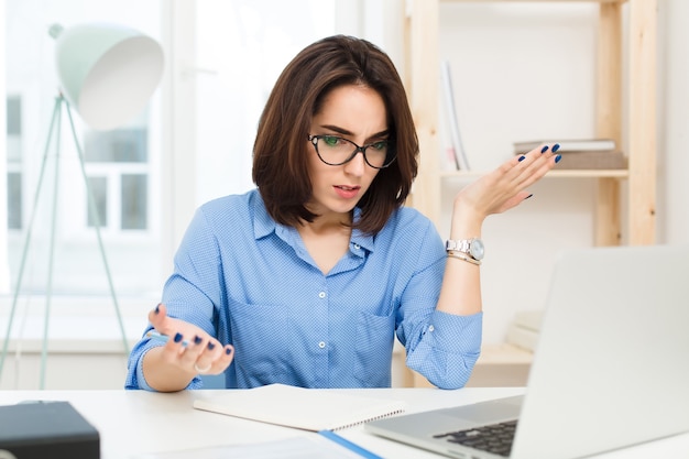 A pretty brunette girl  in blue shirt is sitting at the table in office. She is surprised and  at loss with work.