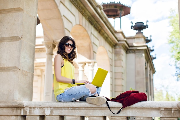 Free photo pretty brunette girl is sitting on fence on old architecture background. she wears yellow shirt, sunglasses, holds yellow laptop on knees, smiling to camera.