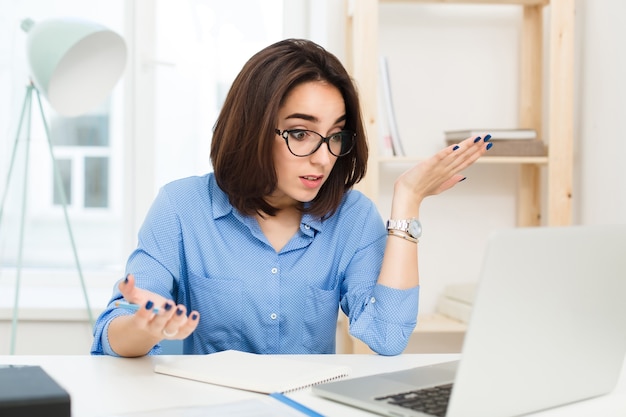 A pretty brunette girl is sitting at the table in office. She wears blue shirt and black glasses. She is at loss with work.