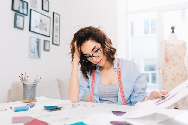 A pretty brunette girl is sitting at the table in the workshop studio. The girl in a blue shirt is busy by looking the sketches in album.