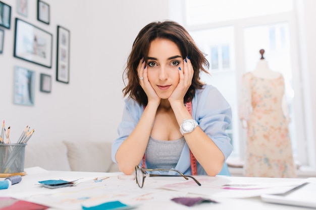 A pretty brunette girl is sitting at the table in the workshop studio. She wears blue shirt and white watch. She supports her face with hands and is looking to camera.