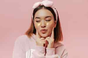 Free photo pretty brunette woman in soft pajamas and headband blows kiss and poses with eyepatches on isolated pink wall