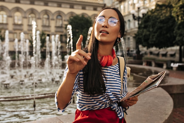 Free photo pretty brunette woman in stylish striped shirt, red headphones and silk skirt points into distance