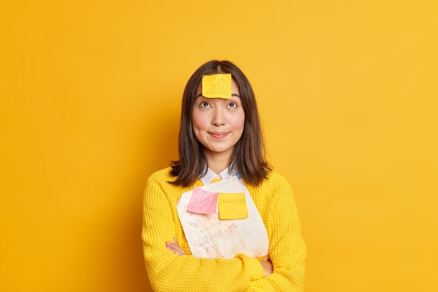 Pretty brunette young Asian female student tries to remember material for exam has deadline memo sticker stuck on forehad concentrated above stands crossed hands