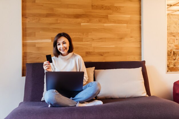 Pretty casual dressed woman is working on laptop computer sitting on dark bed in front of wooden wall at home