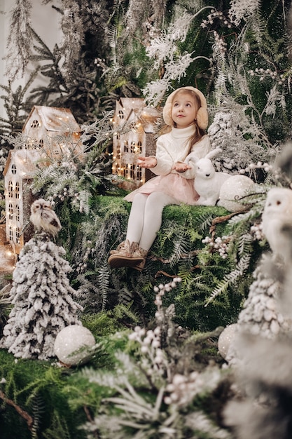 Free photo pretty caucasian child with long fair hair smiles and sits in christmas atmosphere with a lot of decorate trees around her and a little rabbit