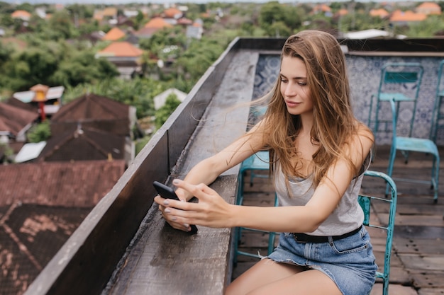 Free photo pretty caucasian girl in denim skirt sitting at roof with smartphone. outdoor portrait of brown-haired lady chilling on street.