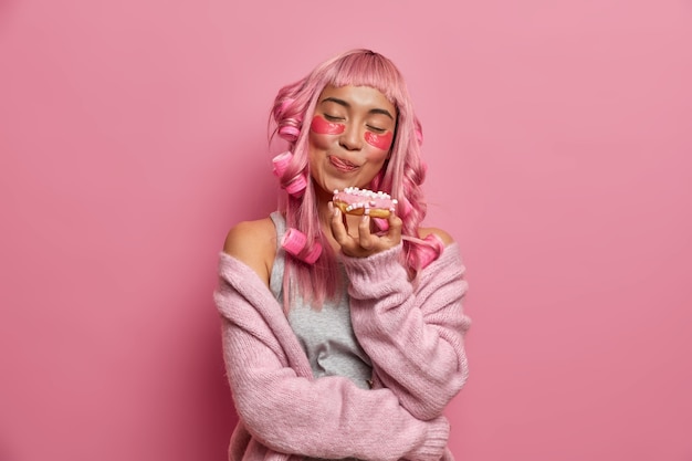 Free Photo pretty charming girl licks lips holds appetizing delicious doughnut applies collagen patches under eyes hair curlers for curly hairstyle warm knitted sweater