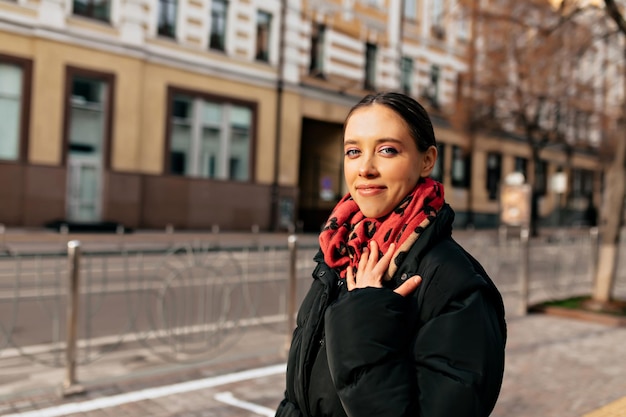 Free photo pretty charming girl with dark hair wearing black jacket and colorful scarf posing at camera with happy smile while walking in the city in sunshine warm sunny winter day in the city