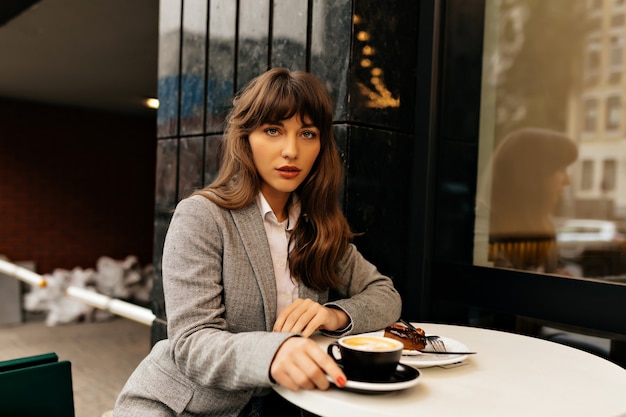 Free Photo pretty charming lady in grey jacket with long wavy hair sitting on city cafeteria is having coffee break