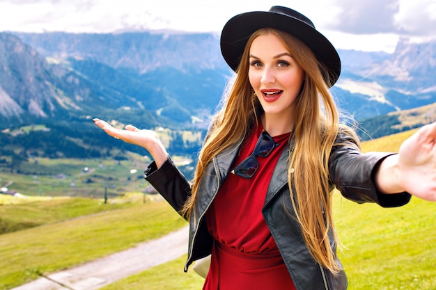 Pretty cheerful young tourist woman wearing stylish leather jacket and trendy hat and showing amazing view on Austrian Alp mountains