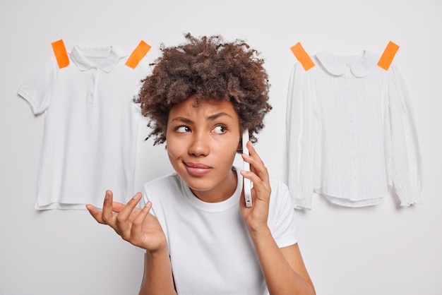 Pretty curly haired woman feels hesitant shrugs shoulder makes telephone call makes decision dressed casually poses against white background with plastered t shirt and blouse So what to do