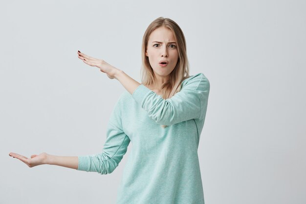 Free photo pretty cute blonde female wearing casual clothes showing something big in size with hands, frowning her brows, pouting lips.