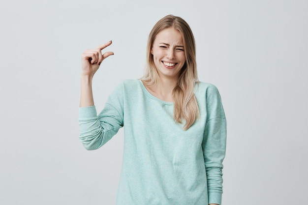 Pretty cute blonde woman in casual clothes showing something small in size with hands while gesturing, smiling broadly with teeth. Blonde European female demonstrating size of little box