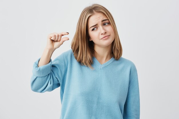 Pretty cute girl wearing casual clothes showing something small in size with hands while gesturing, frowning her brows