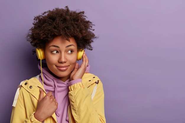 Free photo pretty dark skinned hipster girl listens audio book in headphones, focused aside with dreamy expression, wears casual clothing