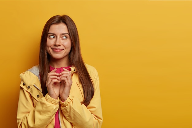 Free photo pretty delighted woman schemes something, steepls fingers and looks with intention aside, has cunning look, made up good idea, smiles pleasantly, wears windbreaker, isolated on yellow  wall