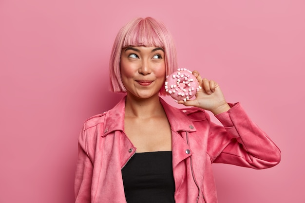 Free Photo pretty dreamy lady with bob dyed hair, holds delicious appetizing donut has temptation to eat high calories dessert, wears stylish pink jacket