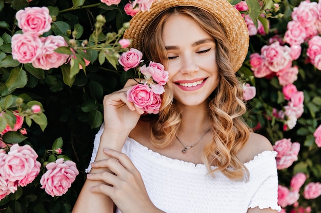 Pretty european girl smiling with eyes closed on nature. Beautiful fair-haired woman enjoying photoshoot with flowers.