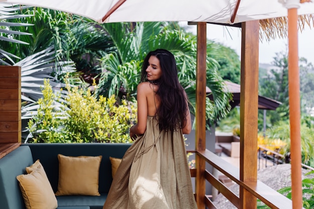 Pretty european positive smiling woman in summer flying dress at natural day light at villa terrace enjoying beautiful vacation, outdoor sofa with pillows on tropical