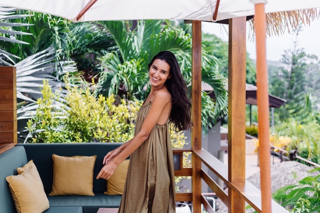 Free Photo pretty european positive smiling woman in summer flying dress at natural day light at villa terrace enjoying beautiful vacation, outdoor sofa with pillows on tropical.