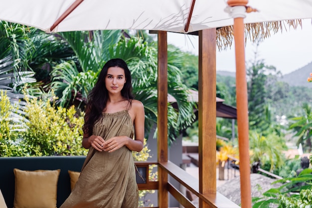 Free Photo pretty european positive smiling woman in summer flying dress at natural day light at villa terrace enjoying beautiful vacation, outdoor sofa with pillows on tropical