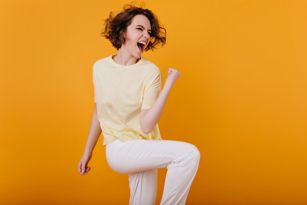 Free photo pretty excited european woman funny dancing  with orange interior. indoor photo of enthusiastic curly girl in white atiire spending time at home.