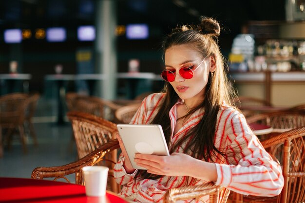 Pretty fashion model in red sunglasses works with her tablet sitting at the cafe 