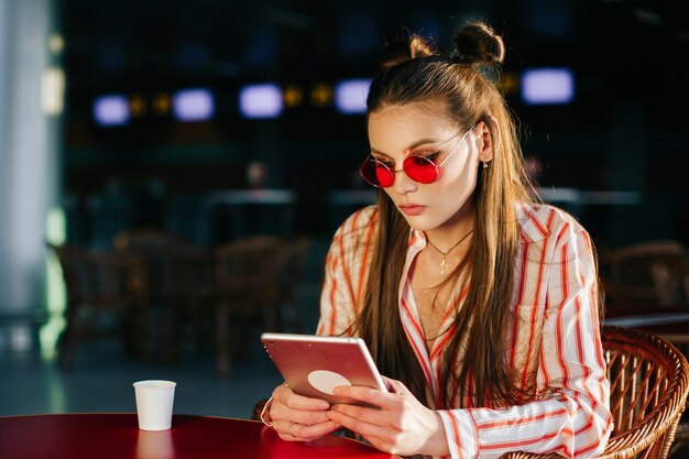 Pretty fashion model in red sunglasses works with her tablet sitting at the cafe 