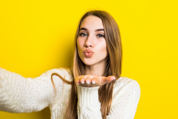 Pretty fashioned girl is making selfie airkiss on her phone wearing wide white sweater on yellow