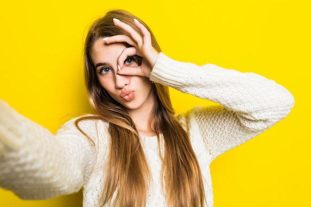 Free photo pretty fashioned girl is making selfie on her phone wearing wide white sweater on yellow