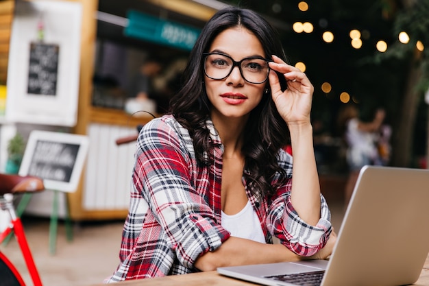 Pretty female freelancer wears trendy glasses posing on blur city. Elegant black-haired girl using laptop in good day.