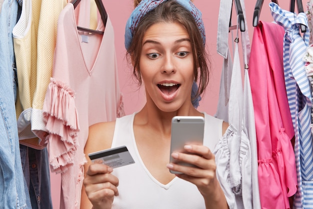 Pretty female standing near wardrobe with clothes, holding smart phone and plastic card, doing shopping online, looking with surprisment in cell phone while having variety of dresses to choose