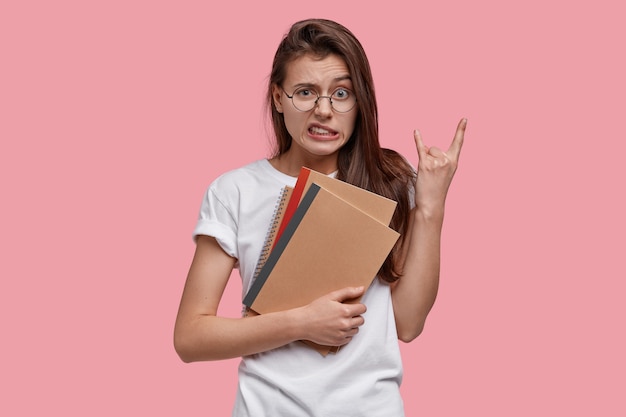 Pretty female with dark hair, makes rock n roll gesture, feels herself cool, carries notepads, dressed in casual white t shirt, models over pink space