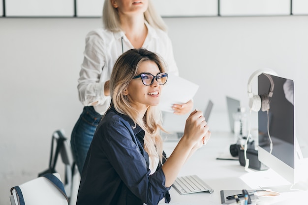 Pretty freelancer girl sitting near computer with black screen and looking away with smile