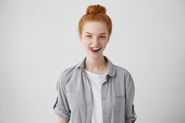 Pretty ginger woman with blue eyes and freckles wearing casual shirt having fun while touching her teeth with tongue having happy expression