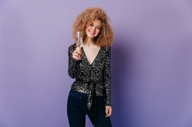 Free photo pretty girl in great mood looks into camera and holds crystal glass of champagne. shot of lady in shiny top on purple space.