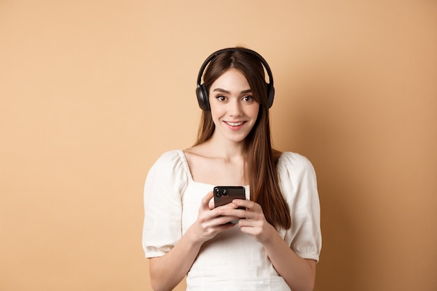 Pretty girl in headphones smiling at camera listening music and using mobile phone app beige background