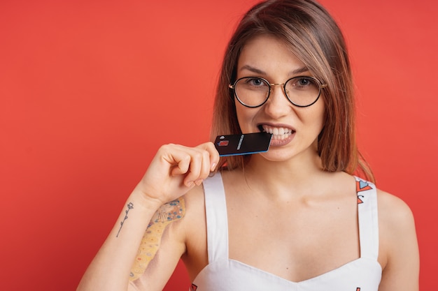 Pretty girl holding with hand a plastic credit card between her teeth on red wall.