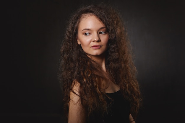 Pretty girl looking sideways, thinking over something. Gorgeous young dark haired Caucasian female with beautiful features having deep in thoughts look posing isolated at black wall