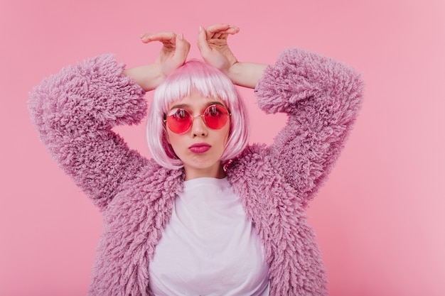 Free photo pretty girl posing in trendy fluffy jacket. jocund european lady in shiny periwig and sunglasses standing on pink wall