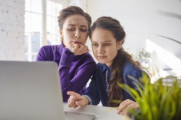 Pretty girl sitting in front of open portable computer at home, pointing finger at screen, mother next to her feeling uncertain and shocked