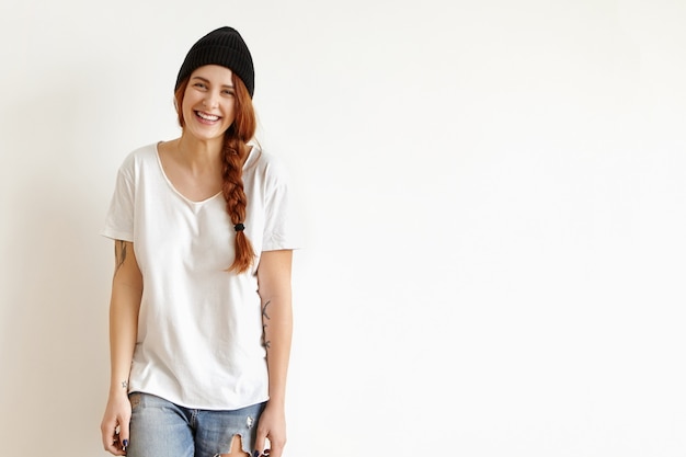 Free photo pretty girl in stylish black hat, white t-shirt and ragged jeans smiling, standing isolated against studio wall