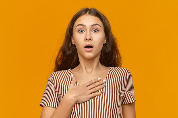 Pretty girl wearing stylish striped summer dress expressing shock and full disbelief keeping hand on her chest