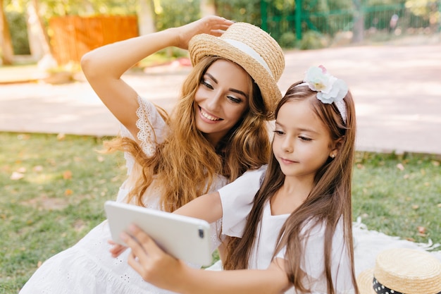 Free photo pretty girl in white dress holding smartphone and making selfie with laughing mom walking down the street. outdoor portrait of glad young woman in hat posing while brunette daughter taking photo.