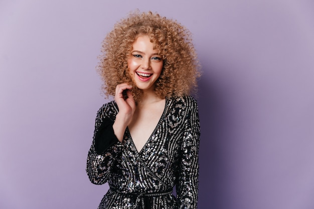 Pretty girl with blond curls cute smiles. Snapshot of lady in shiny v-neck blouse on purple space.